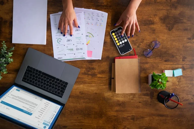 Overhead view of a person using a phone calculator while working on financial charts next to a laptop