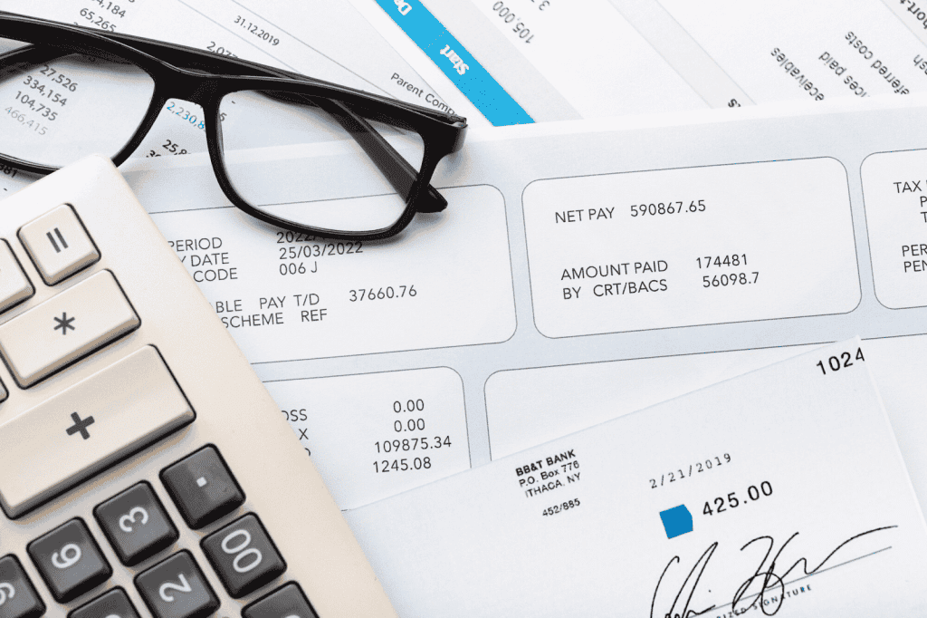 A close-up of a desk with a calculator, a pair of glasses, and financial documents, including a payslip and a bank envelope