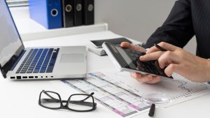 Business professional calculating financial figures at an office desk with laptop and reports.