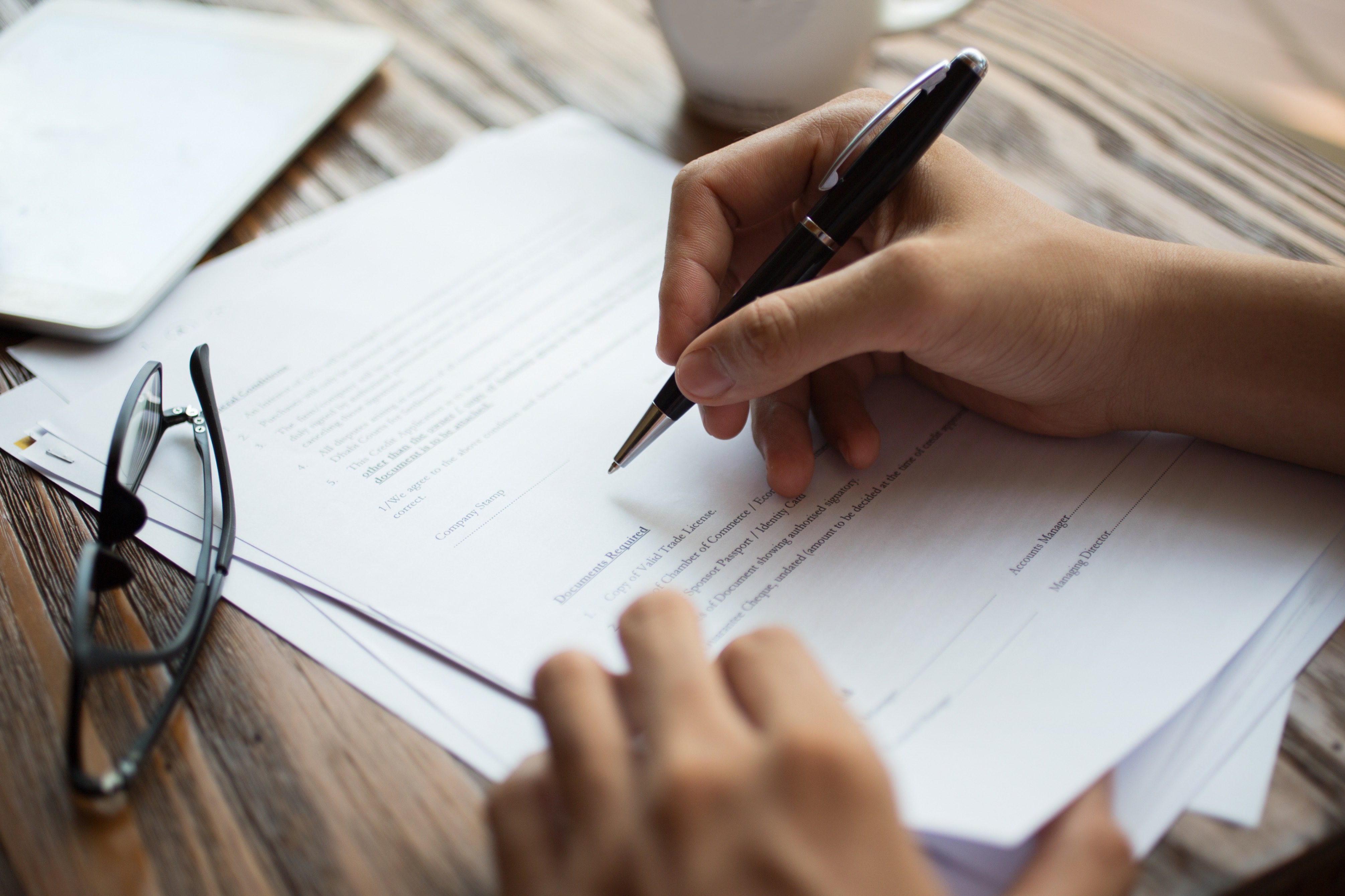 Person signing a document with a pen on a wooden desk.