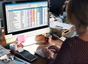 Person typing at a desk with a spreadsheet on screen and cluttered workspace. 