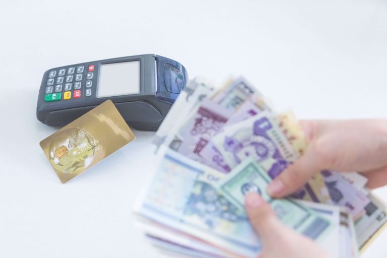 Close-up of a payment terminal with a gold credit card and hand holding colorful banknotes, symbolising cash vs card payment options.