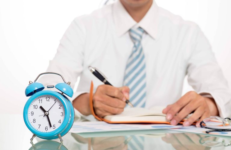 man writing in a diary on a table with a clock on it