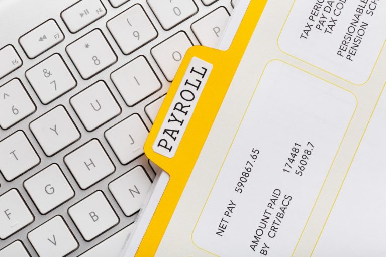 A magnifying glass is held over a gray stone surface, perfectly centering and enlarging the word "TAX" spelled out on small wooden tiles