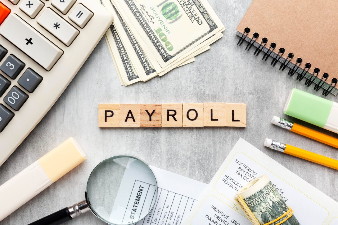 Payroll concept with calculator, US dollar cash, payslip, magnifying glass, stationery, and wooden blocks spelling “PAYROLL” on a desk.